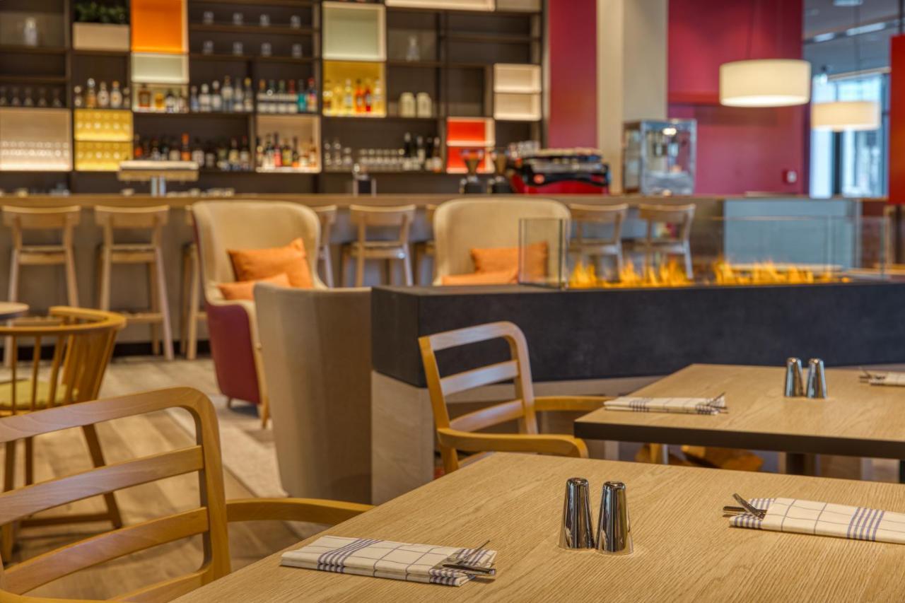 Intercityhotel Berlin Airport Ber Terminal 1 & 2 Schönefeld Εξωτερικό φωτογραφία The photo shows a modern restaurant or bar interior. In the foreground, there are wooden tables set with napkins and condiments. The seating includes wooden chairs and plush seating in various colors in the background. A stylish bar area is visible w